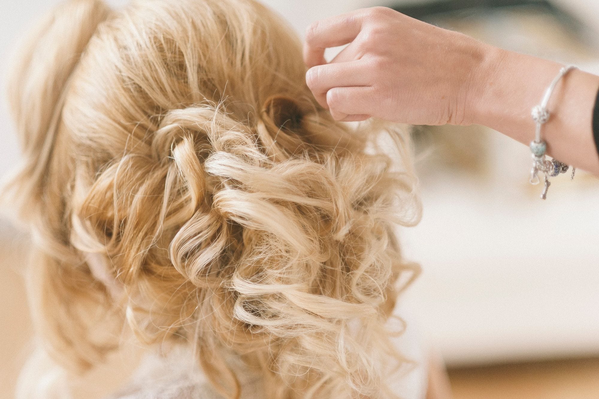 A hairstylist arranging a blonde bridal updo, seen from behind.